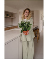 Een glimlachende vrouw met krullend blond haar, gekleed in een lichtgroen pak, staat in een moderne keuken en houdt een Kunstbloem Kalanchoë plant UV 31 cm wit met delicate witte bloemen vast.