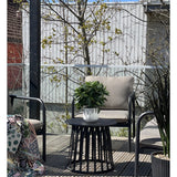 Een modern terras met twee stoelen met kussens, een ronde zwarte tafel met de Kunstplant Laurier UV d33 cm als middelpunt en een deken met patroon op één stoel. Bomen en een houten hek verschijnen op de achtergrond onder een heldere hemel.