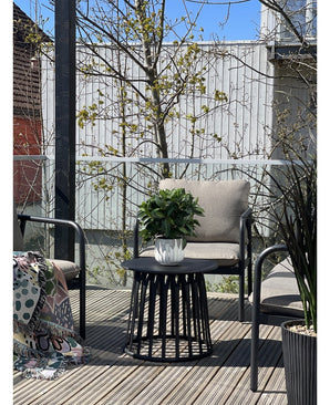 Een gezellig terras met twee stoelen met kussens, een ronde zwarte tafel met de Kunstplant Laurier d33 cm UV en een deken met patroon gedrapeerd over één stoel. Bomen en een houten hek zijn zichtbaar op de achtergrond.