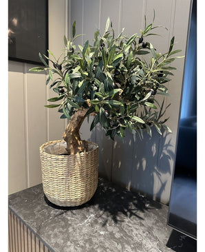 De Kunst Olijfmontage UV 60cm, met groene bladeren en zwarte olijven, zit in een geweven mand op een donker marmeren oppervlak tegen een lichte wand met panelen -onderhoudsvriendelijk en UV-bestendig voor langdurige schoonheid.