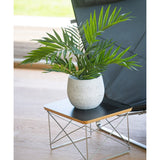 Een Kunstpalm Parlour UV 60 cm staat in een pot op een klein modern bijzettafeltje met een zwart blad en metalen draadpoten, naast een zwarte stoel op een lichte houten vloer.