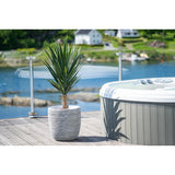 Een Kunstplant Yucca 100 cm UV staat op een houten dek naast een hot tub met glazen reling, met uitzicht op een rustig meer en groene heuvels bezaaid met huizen.