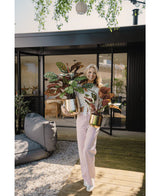Een vrouw staat buiten op een houten terras, glimlacht en houdt twee Kunstplant Calathea Makoyana 60 cm rood in pot vast. Ze draagt een lichtroze broek en een luchtig topje. Achter haar staat een modern glazen gebouw met terrasmeubilair.