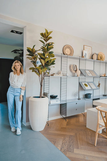 Een vrouw in een witte blouse en lichtblauwe broek staat naast een Kunstplant Yulaniaboom 180 cm in een lichte moderne woonruimte met houten vloeren en open rekken met boeken, kunst en keramiek.
