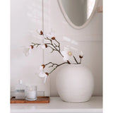 Een Kunstmagnoliatak Real Touch Wit 98cm staat in een ronde witte vaas op een wit oppervlak naast een brandende glazen kaars en een klein flesje op een houten dienblad, in een lichte, minimale kamer met een ronde spiegel erboven.