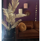 Een moderne woonkamer met een bruin fluwelen bank, rond plooikussen en de Kunstpalm Areca goud 120 cm in een zwarte vaas als gouddecor. Geometrische wandkunst hangt op een donkerpaarse muur, met houten vloeren en verticale lamellen in het zicht.