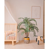 Een gezellige kinderkamer met een licht houten ledikant, de Kunstpalm Cycas Classic 135 cm in een gele plantenbak, een houten speelgoedfiets met het label MIA, minimalistische lijnkunst aan de muur, lichthouten vloeren en zachtroze beddengoed voor extra warmte.