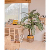 In een gezellige, lichte kamer staan de Kunstpalm Cycas Classic 135 cm in een stijlvolle plantenbak, een rieten speelgoedkar, houten meubels en kinderboeken bij een raam met een bamboe rolgordijn.