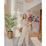 Een glimlachende vrouw in lichtbeige kleding en sneakers staat in een lichte, moderne woonkamer met houten meubels en ingelijste kunst, en houdt een kleurrijk boeket vast. De Kunstpalm Kentia 135 cm voegt een tropisch tintje toe aan de ruimte.