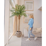 Een vrouw in een blauwe blouse en spijkerbroek loopt langs de Kunstpalm Phoenix De Luxe 210 cm bij een groot raam in een lichte, moderne woonkamer met beige muren, een bank en ingelijste kunst aan de muur.