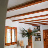 Een gezellige woonkamer met witte muren, houten balken, een witte sectionele bank, de Kunstpalm Phoenix De Luxe 210 cm, houten raam en deur, ingebouwde planken met manden en aardewerk, en natuurlijk licht dat door het raam naar binnen valt.