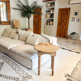 Een gezellige woonkamer met een witte bank, ronde houten bijzettafel, Kunstpalm Phoenix De Luxe 210 cm, gevlochten manden op planken, houten deuren, een ronde spiegel en neutraal getinte vloerkleden op een lichte vloer.