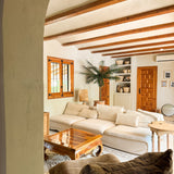 Een gezellige woonkamer heeft een grote crèmekleurige sofa, houten salontafel, Kunstpalm Phoenix De Luxe 210 cm in een pot, houten balkenplafond, ingebouwde planken met aardewerk, natuurlijk raamlicht en een warme, rustieke inrichting.