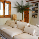 Een gezellige woonkamer met een grote crèmekleurige bank, de Kunstpalm Phoenix De Luxe 210 cm in de hoek, een houten lamp, ingebouwde planken met manden, houten plafondbalken en een raam met houten omlijsting.