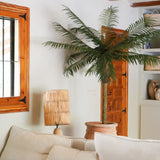 Een gezellige woonkamer met een witte bank, een rotan tafellamp, de Kunstpalm Phoenix De Luxe 210 cm in een pot, houten balken aan het plafond, een raam met houten kozijnen en een houten deur op de achtergrond.