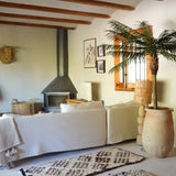 Een gezellige woonkamer heeft een witte bank, een vloerkleed met patroon, een houtkachel, ingelijste kunst en houten balken. Zonlicht stroomt naar binnen vanuit een raam naast de weelderige Kunstpalm Phoenix De Luxe 210 cm, waardoor de ruimte tot leven komt.
