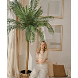 Een glimlachende vrouw staat naast de Kunstpalm Phoenix De Luxe 250 cm in een lichte, stijlvolle woonkamer met neutrale inrichting en grote abstracte kunst. Ze houdt een plantje vast en leunt nonchalant tegen een beige bank met kussens.