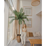 Een vrouw in lichtgekleurde kleding staat naast de Kunstpalm Phoenix De Luxe 250 cm in een lichte, moderne woonkamer met grote ramen, een beige bank, rieten lamp, houten salontafel en ingelijste muurkunst.