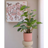 De Kunstplant Appelblad 90 cm, een groenbladige potplant in een roze geribbelde pot, staat op een beige sokkel voor een muur met abstracte roze bloemsierkunst en een doorschijnend wit gordijn aan de rechterkant.