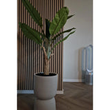 De Kunstplant Bananenboom 150 cm met grote groene bladeren staat op een houten vloer voor een houten wand met latten. Rechts staat een moderne bladloze ventilator in de kamer met neutrale muren en vloer.