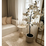 Een gezellige woonkamerhoek met een witte bank, beige kussens, een kleine ronde bijzettafel met gouden kandelaars, transparante gordijnen en de Kunstplant Bougainvillea 120 cm wit voor een elegante touch.