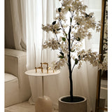 Een Kunstplant Bougainvillea van 120 cm groot staat bij een goudomrande spiegel naast een kleine ronde witte bijzettafel met gouden kandelaars, hoge witte kaarsen, transparante witte gordijnen en een crèmekleurige bank in een lichte kamer.