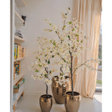Een Kunstplant Bougainvillea 120 cm groot met witte bloesems in een goudkleurige pot met textuur staat bij een raam met transparante gordijnen, naast boekenplanken vol boeken en decor in een lichte, moderne kamer.