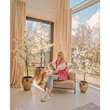 Een glimlachende vrouw zit in een beige fauteuil bij grote ramen met transparante gordijnen, omringd door Kunstplant Bougainvillea 120 cm wit in gouden plantenbakken. Het zonlicht valt naar binnen en verlicht de moderne, gezellige kamer.