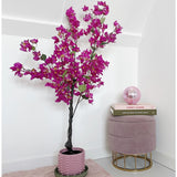De Kunstplant Bougainvillea 125 cm Paars met levendige paarse bloemen staat naast een roze fluwelen kruk met daarop gestapelde boeken en een roze glazen bol in een minimale, moderne kamer met witte muren.