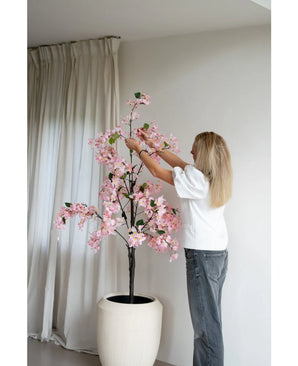 Een persoon met lang blond haar schikt de Kunstplant Bougainvillea 150 cm roze in een grote witte pot bij een raam met vloerlange witte gordijnen in een minimalistische kamer.