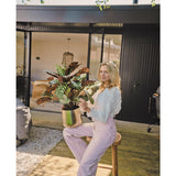 Een blonde vrouw in een witte blouse en lichtpaarse broek zit buiten op een krukje met de Kunstplant Calathea Makoyana UV 75 cm rode potplant, op een houten vlonder voor een modern huis met glazen deuren.