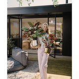 Een vrouw staat op een houten dek voor een modern huis, glimlacht en houdt twee Kunstplant Calathea Makoyana UV 75 cm rode potplanten vast. Ze draagt een lichtroze broek en een wit topje. Buiten zitplaatsen en groen op de achtergrond.