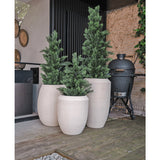 Drie hoge, witte plantenbakken met textuur en Kunstplant Conifeer UV 120cm kunstgroene planten staan op een houten terras naast een modern huis. Een zwarte buitengrill staat vlakbij, met een stenen muur en groen op de achtergrond.