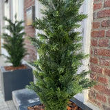 Een close-up van de Kunstplant Conifeer UV 120cm, een groene conifeer kunstplant in een rechthoekige zwarte plantenbak met bruine schorsmulch, uitgestald op een stoep bij een bakstenen gebouw; een andere dergelijke plantenbak staat op de achtergrond.