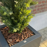 Een close-up van de Kunstplant Conifeer UV 120cm, een realistische groene conifeer in een moderne vierkante grijze plantenbak met bruine schorssnippers, uitgestald tegen een bakstenen muur en betegelde grond.
