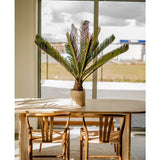 Een houten eettafel met twee bijpassende stoelen staat bij een groot raam. Op de tafel staat een Kunstplant Cycus Palm Deluxe 95 cm en het zonlicht stroomt naar binnen en werpt zijn schaduwen op het meubilair en de vloer.