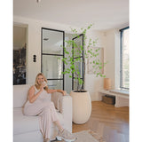 Een vrouw in lichte loungekleding zit op een beige bank en glimlacht met een kopje koffie in een moderne woonkamer met houten vloeren, ramen van vloer tot plafond en de Kunstplant Enkianthus 180 cm als grote potplant.