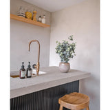 Een modern aanrecht heeft een messing kraan, zeepdispensers, de Kunstplant Eucalyptus 61 cm potplant en twee houten krukjes. Boven het aanrecht houden planken op een lichte muur potten en flessen.