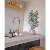 Een modern aanrecht heeft een gouden kraan, drie zeepdispensers, de Kunstplant Eucalyptus 91 cm en houten krukjes. Minimalistische planken boven tonen potten met verschillende ingrediënten voor een gezellige uitstraling.
