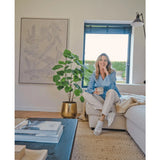 Een vrouw zit op een beige bank in een gezellige, moderne woonkamer, met een mok in de hand en een glimlach. De ruimte wordt gekenmerkt door de Kunstplant Ficus 120 cm in een gouden pot, abstracte kunstwerken en zonlicht dat door blauwe jaloezieën naar binnen schijnt.