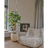 Een gezellige, beige loungestoel met structuur staat naast de Kunstplant Ficus 150 cm in een moderne woonkamer met lichte houten vloeren, neutrale muren, een raam en vloerlange grijze gordijnen.