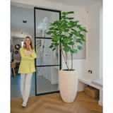 Een vrouw in een gele blazer en witte broek glimlacht naast de Kunstplant Ficus 180 cm in een moderne, lichte kamer met houten vloer en minimalistisch decor.