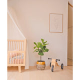 Een minimale kinderkamer met een licht houten ledikant, een doorschijnende luifel, Kunstplant Ficus Tabaksplant Deluxe 90 cm, houten speelgoed met het label Mia en een minimalistische lijnkunst ingelijste print op een effen witte muur.