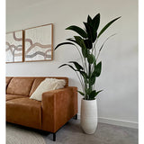 Een moderne woonkamer met een bruine leren bank, gestreepte kussens en de Kunstplant Heliconia 180 cm in een witte vaas. Daarboven hangt abstracte kunst aan een witte muur met een grijze vloer en een beige vloerkleed - ideaal voor kunstplant kopen inspiratie.