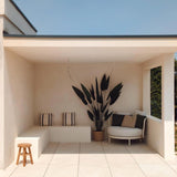 Een moderne, minimalistische patio met crèmekleurige tegels, een ingebouwde bank met gestreepte kussens, een Kunstplant Heliconia 210 cm voor een luxe touch, een klein houten krukje en een ronde stoel met kussens onder een plat dak tegen een helderblauwe hemel.