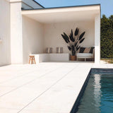 Modern buitenzwembad met lichte tegelvloer, een witte overdekte zithoek met gestreepte kussens, een Kunstplant Heliconia 210 cm voor een luxe touch, een klein krukje, een sofa, blauwe lucht erboven en een groene haag om het plaatje compleet te maken.