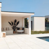 Een modern buitenterras met witte tegelvloeren, schaduwrijke zitplaatsen met een luxe uitstraling, de Kunstplant Heliconia 210 cm als middelpunt, gezellige kussens, een houten kruk en een zwembad op de voorgrond onder een strakblauwe hemel.