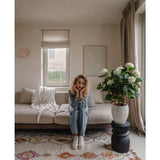 Een vrouw met krullend blond haar zit op een beige bank in een lichte woonkamer en laat haar kin op haar handen rusten. Naast haar staat de Kunstplant Hortensia UV 85 cm groot, verlicht door licht door een raam met beige gordijnen.