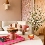 In een gezellige woonkamer staat een witte salontafel met een roze boek, schaal, sokkelschaal, kaars en de Kunstplant Kersenbloesem 120 cm crème. Op de achtergrond staan een beige bank met gestreepte kussens en roze behang met patroon.