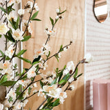 Een Kunstplant Kersenbloesem 120 cm crème met witte en lichtroze bloesems staat voor een houten deur. Daarachter zijn een roze kruk, spiegel en roze behang met patroon te zien.